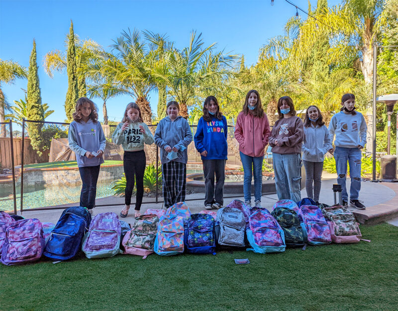 Young girls posing behind a line of backpacks.