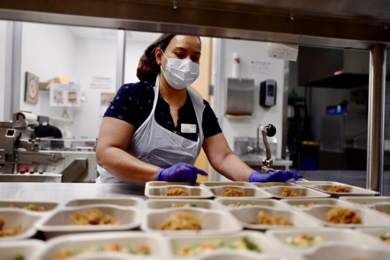 Cook, wearing a mask, laying out trays containing food.