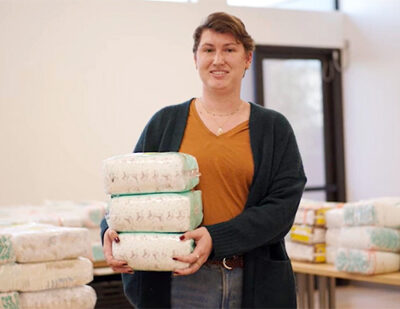 A diaper bank volunteer holds packages of diapers while standing in front of tables stacked with diaper donations.