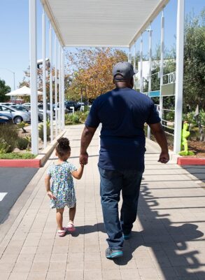 a father walking down the sidewalk while holding his little girl's hand