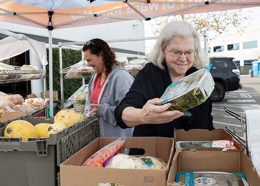Two women sorting groceries under a tent. One woman holds a container of greens. Boxes and bins filled with produce and packaged food are visible.
