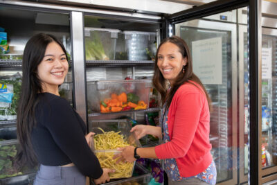 Two women are standing in front of a refrigerator filled with vegetables in the JFS Corner Market, smiling while placing or removing containers of produce.