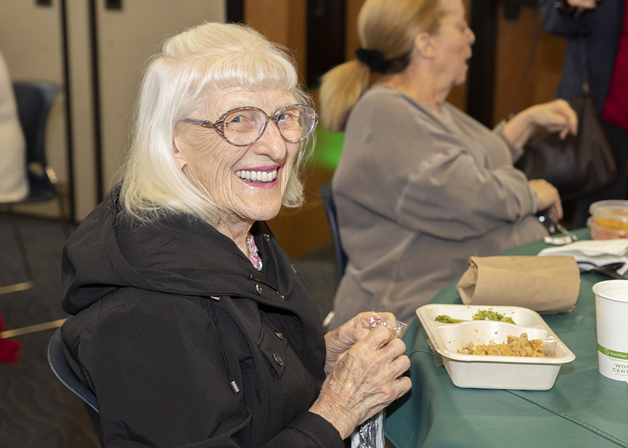 An older woman with glasses and white hair smiles while sitting at a table with a meal, alongside another woman in a casual setting.