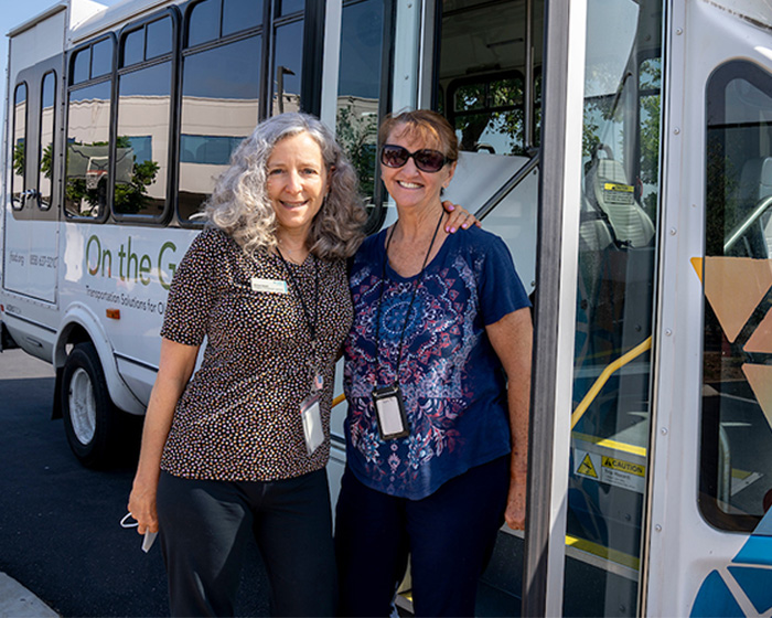 Two women stand beside a shuttle bus, smiling broadly. Behind them, is the On the Go bus opening its doors.