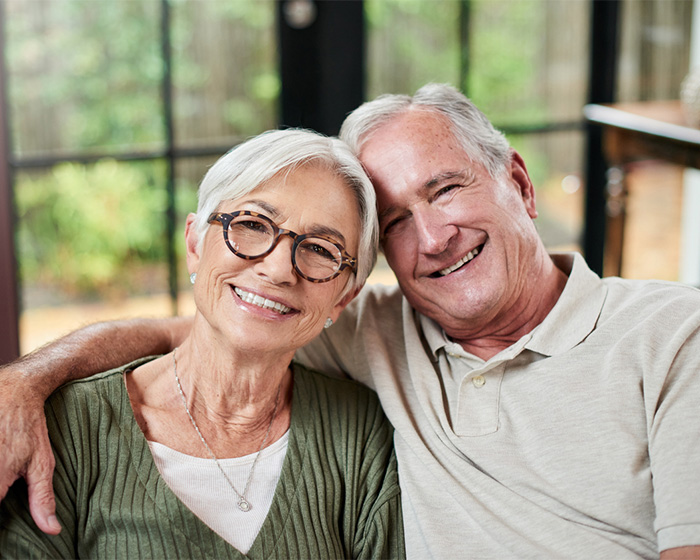 An elderly couple, embodying the spirit of giving, sits closely together, smiling warmly at the camera. The man wears a light polo shirt, while the woman dons a green sweater with glasses. They are inside, with a blurred window view of greenery behind them.