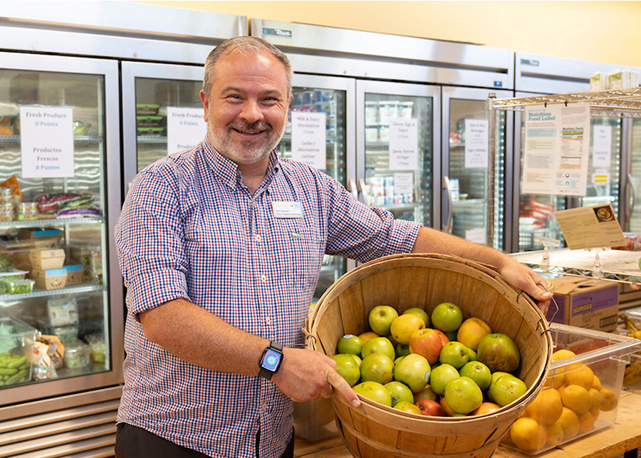A person smiling and holding a basket of green and red apples in the Corner Market with refrigerators in the background.