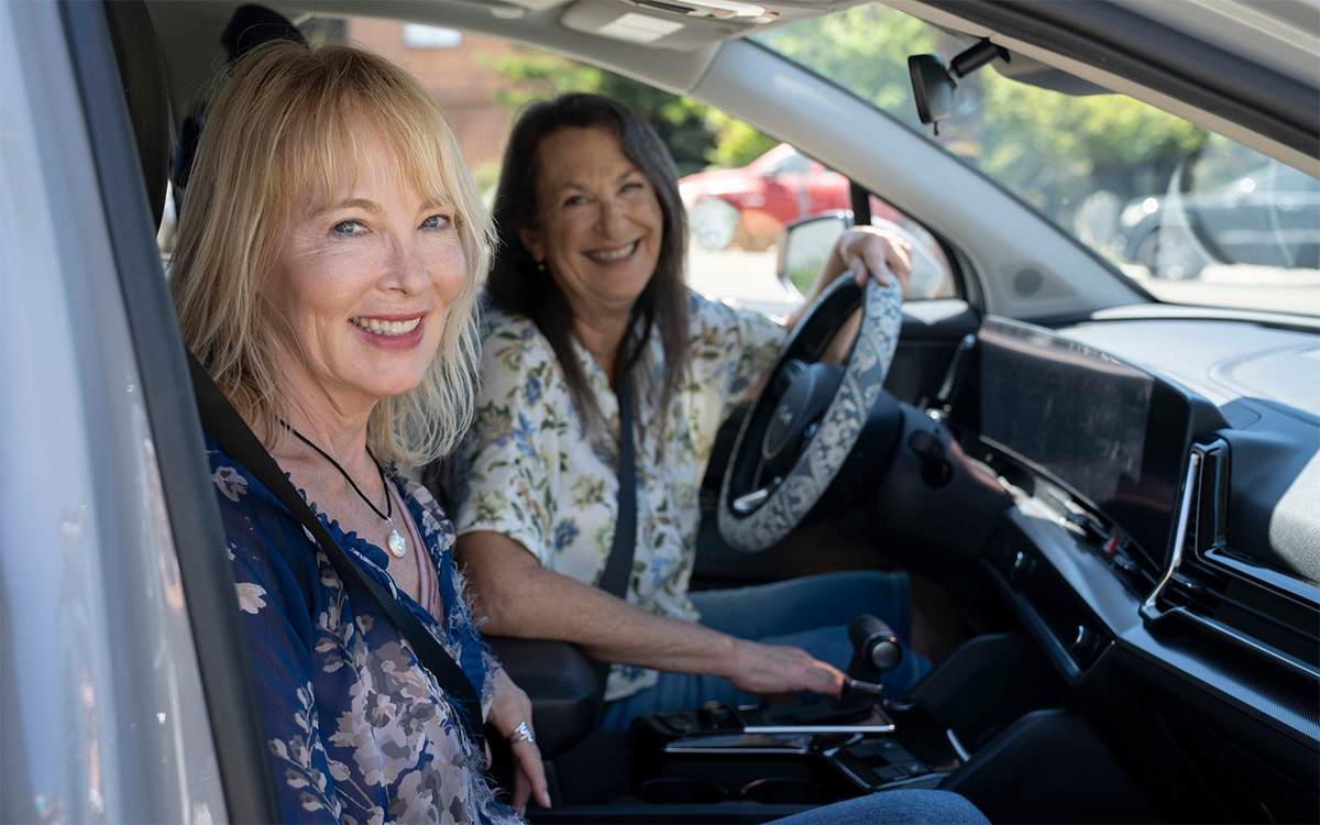 Two smiling women sitting in the front seats of a car, one holding the steering wheel